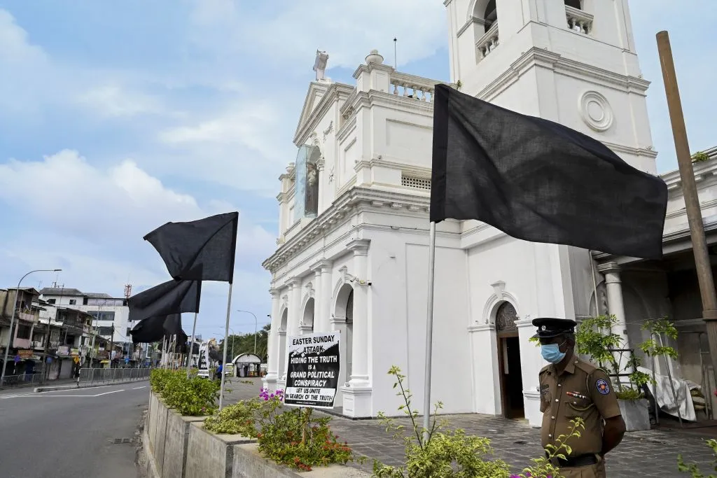 A policeman stands guard at St. Anthony's church in Colombo, Aug. 21, 2021, next to a placard and black flags placed in protest for the alleged failure to prosecute those responsible for the bomb attacks of Easter Sunday 2019. Credit: Ishara S. Kodikara/AFP via Getty Images.?w=200&h=150